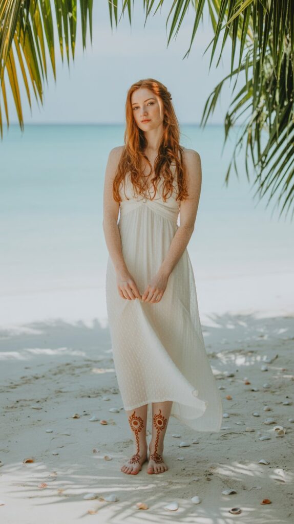 Redhead bride on a beach, ivory maxi dress, barefoot with crescent moon and sun-themed henna across tops of feet, shells and palm fronds around her.
